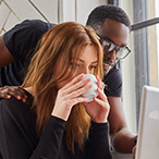 Woman drinking from a mug and looking intently at something with man peering over her shoulder