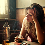 Woman sitting at a table, staring into space, with drink in hand and bottle of alcohol next to her